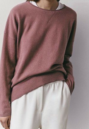 Een persoon draagt een lang roze trui met stof over een wit overhemd, gecombineerd met loszittende witte broek, hand in de zak, neutrale achtergrond.