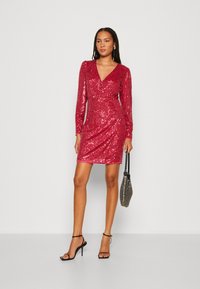 Robe rouge à paillettes avec des manches longues, un décolleté en V et une silhouette ajustée. Associée à un sac à main métallique et des talons noirs à lanières.