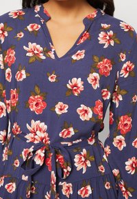 Marinblå blommig klänning med röda och vita blommor, V-ringning och långa ärmar. Har ett knytbälte och ett utsvängd kjol. Mjuk, texturerad tyg.