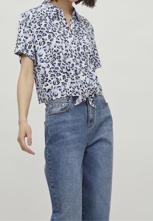 Femme portant une chemise à manches courtes boutonnée avec imprimé léopard nouée à la taille et un jean en denim bleu taille haute sur un fond uni.