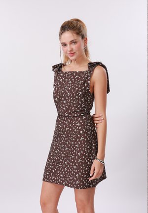 Mlada ženska s svetlo blond lasmi v rjavi obleki brez rokavov z drobnim belim cvetličnim vzorcem, ki pozira na svetlem ozadju.