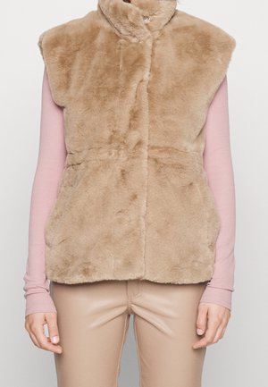Beżowa kamizelka bez rękawów z beżowego futerka sztucznego z wysokim kołnierzem, noszona na różowej bluzce z długim rękawem i beżowych spodniach, ręce swobodnie opadające wzdłuż ciała.