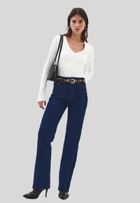 Mladá žena v bílém dlouhém rukávu, tmavě modrých džínech s vysokým pasem a leopardím páskem, černých lodičkách a nese černou kabelku přes rameno.