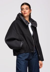 Femme portant une veste noire zippée surdimensionnée avec un col rembourré haut et un jean bleu, regardant vers sa gauche sur un fond uni.