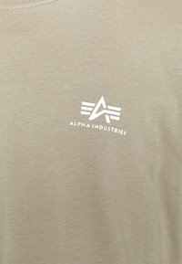 Béžová tkanina s bílým logem Alpha Industries, které obsahuje stylizované "A" a tři vodorovné pruhy nad textem.