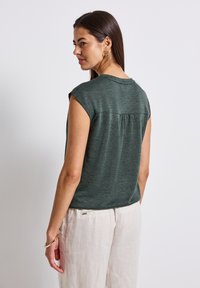 Green short-sleeve top with a round neckline, featuring a back pleat detail and soft, textured fabric. Pairing with light-colored linen pants.