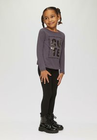 Lila Langarmshirt mit schwarzem Metallic-Text, kombiniert mit schwarzen Leggings und glänzenden Schnürstiefeln. Glatte Stoffstruktur.