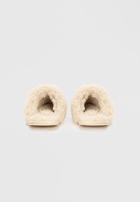 Cremefarvede tøfler med en fluffy tekstur, åben rygdesign og en flad elfenbensfarvet sål. Foret med blødt stof for komfort.