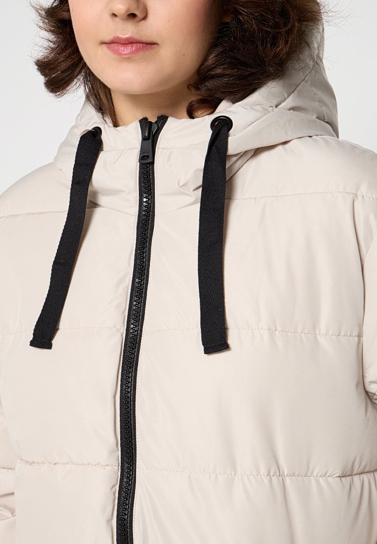 Chaqueta acolchada de color beige claro con capucha, que presenta una cremallera central y cordones negros. Textura suave y detalles de diseño acolchado.