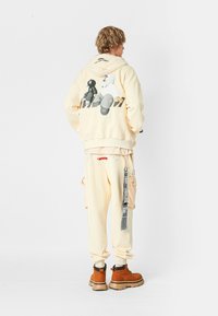 Joven de pie, vistiendo una sudadera beige con personajes de dibujos animados en la parte de atrás, pantalones a juego con bolsillos laterales y correas, y botas marrones.