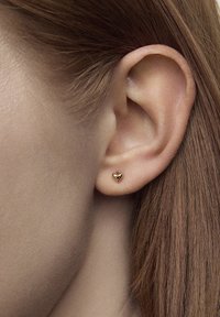 Złota kolczyk w kształcie serca przymocowany do ucha modelki, charakteryzujący się gładką teksturą i subtelnym połyskiem, podkreślający kontury ucha.