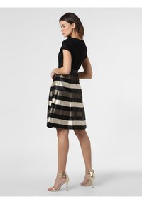 Schwarzes Oberteil mit figurbetontem Design, kombiniert mit einem knielangen Rock in abwechselnden schwarzen, goldenen und metallischen Streifen. Goldene, schnürbare hohe Absätze.