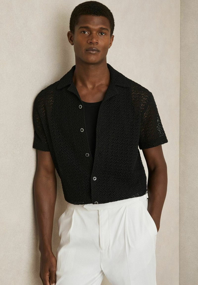 Cămașă scurtă din tricot negru cu un design cu nasturi și textura țesută deschisă, asociată cu pantaloni albi cu talie înaltă și pliuri.