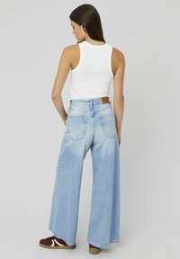 Femme aux longs cheveux bruns portant un haut sans manches blanc, un jean bleu clair à jambes larges et des baskets bordeaux, se tenant de dos.