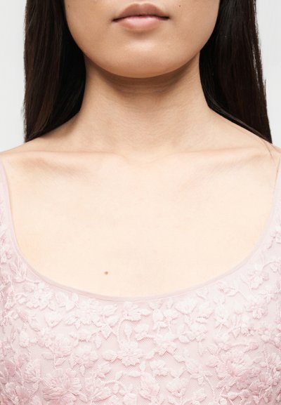 Το κάτω μέρος του προσώπου και ο λαιμός μιας γυναίκας με ένα μικρό ελιές, που φοράει μια ανοιχτό ροζ φλοράλ δαντελένια μπλούζα με λεπτές τιράντες.
