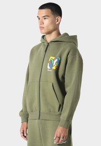 Olivegrøn zip-up hoodie lavet af blødt stof, med en frontlomme og et farverigt abstrakt design på venstre bryst.