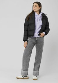 Veste matelassée noire au design ajusté, sweat à capuche lavande en dessous, jean gris à jambes larges et baskets blanches avec des accents noirs.