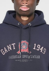 Hanorac bleumarin din bumbac, cu un logo grafic în roșu și alb. Include șnururi cu capete metalice, manșete și tiv cu model rib.