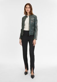 Grüne Leder-Motorradjacke mit Seitenstreifen, eng anliegendes weißes Top, schwarze maßgeschneiderte Hose mit Schlitzen und schwarze spitze Schuhe.