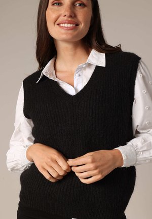 Pull sans manches noir en maille avec un col en V, présentant un motif côtelé texturé. Porté par-dessus une chemise blanche boutonnée.