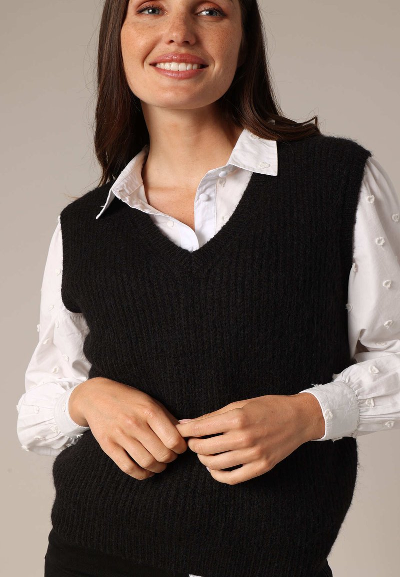 Pull sans manches noir en maille avec un col en V, présentant un motif côtelé texturé. Porté par-dessus une chemise blanche boutonnée.