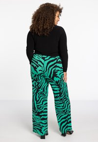 Hoge taille, wijde broek in een groen en zwart zebra print, gemaakt van een lichte stof. Gestyled met een aansluitend zwart topje en blokhakschoenen.