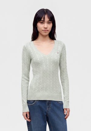 Femme portant un pull gris clair en tricot torsadé à col en V et un jean bleu, debout devant un fond blanc uni.