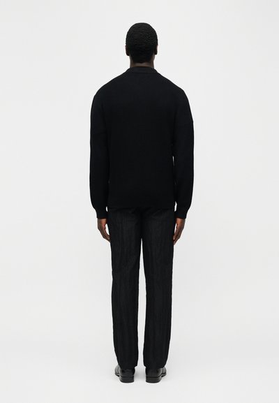 Άνδρας που στέκεται με την πλάτη, φοράει μαύρο πουλόβερ, μαύρο υφασμάτινο παντελόνι και μαύρα παπούτσια σε απλό ανοιχτό φόντο.