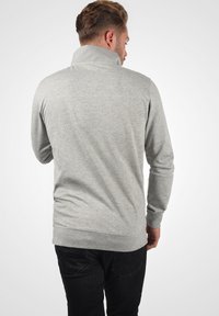 Grå strikka genser med høy krage og lange ermer, som har en jevn tekstur og ribbede mansjetter og kant. Sorte jeans fullfører antrekket.