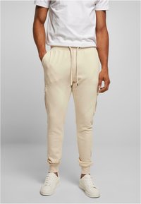 Beige cotton joggers with side pockets, elastic waistband, and cuffed ankles, paired with white sneakers and a plain white T-shirt.