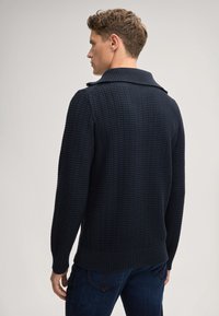 Dunkelblauer Strickpullover mit strukturiertem Muster, Schalkragen und gerippten Bündchen. Über dunkelblauen Jeans getragen. Rückansicht gezeigt.