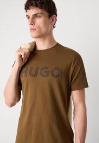 Barna pamut póló kerek nyakkal, amelyen nagy, sötétbarna "HUGO" logó látható elöl, egy modellel, aki egy bézs zakót lógat a vállára.