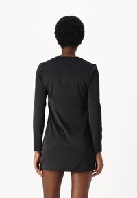 Svart långärmad klänning av mjukt material, med en enkel design och minimalistisk detalj på ryggen. Kort i längd med rak skärning.