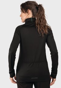 Schwarzes Langarmshirt mit hohem Kragen, featuring eine strukturierte Oberfläche, grüne Nähdetails und einen abgerundeten Saum mit einem Logo auf der Rückseite.