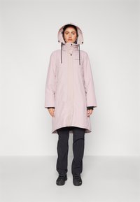 Giacca impermeabile rosa chiaro con cappuccio, caratterizzata da cordini neri e chiusura a zip, indossata sopra pantaloni e scarpe scuri.
