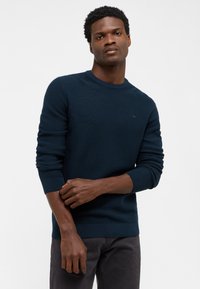 Mörkblå, ribbad stickad tröja med rund hals och långa ärmar; har en subtil logotyp på bröstet och en texturerad tyg.