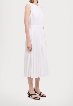 Weißes ärmelloses Kleid mit tailliertem Oberteil und plisseeartigem Rock, der unter das Knie fällt, kombiniert mit schwarzen Riemchensandalen.