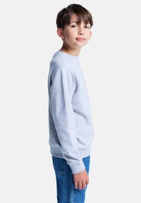 Un ragazzo sta in piedi di lato, indossando una felpa grigia e jeans blu, con un'espressione neutra, contro uno sfondo bianco e semplice.