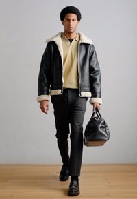 Μαύρο δερμάτινο bomber μπουφάν με κολάρο από shearling, μπεζ πόλο μπλουζάκι, σκούρο παντελόνι, μαύρα loafers και υφασμένο μαύρο duffel τσάντα.