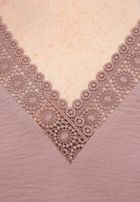 Ροζ τοπ με V-λαιμόκοψη διακοσμημένη με περίτεχνη δαντέλα που παρουσιάζει κυκλικά σχέδια και υφή υφάσματος.