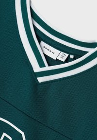Camisa de manga corta verde oscuro con un cuello en V de rayas blancas, fabricada en un tejido transpirable. Presenta una etiqueta con el nombre de la marca y detalles del tamaño.