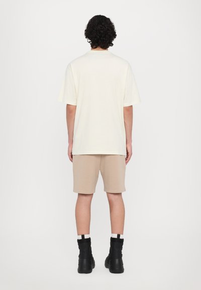 Helles gelbes T-Shirt mit kurzen Ärmeln, kombiniert mit beigen Shorts. Model trägt schwarze Stiefel und steht vor einem schlichten weißen Hintergrund.