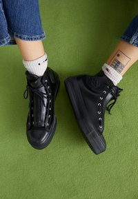 Basket high-top noires en matériau brillant avec des œillets en métal, des lacets plats et des semelles texturées. Portées avec des chaussettes blanches à motifs.