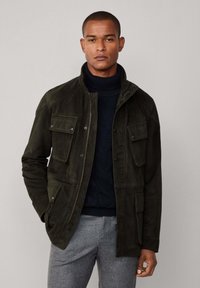Giacca in suede verde oliva con quattro tasche anteriori, chiusura a scatto e cerniera, colletto alto e tessuto texture, abbinata a un maglione blu navy.