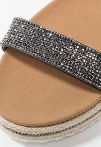 Sandale à slide avec un semelle en daim brun, une lanière en strass noir scintillant et une semelle en jute. Pas de marque visible.