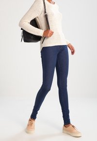 Personne portant un pull en maille crème, un jean skinny bleu foncé, des baskets pêche, et portant un sac à main noir sur l'épaule.