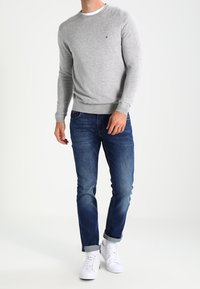 Pull gris à col rond en tissu doux, associé à un jean bleu foncé. Des baskets blanches complètent la tenue, avec les pans du pantalon retroussés.
