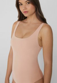 Mladá žena v růžovém upnutém body bez rukávů, zobrazená od poloviny stehna k těsně nad hlavou, na jednobarevném pozadí.
