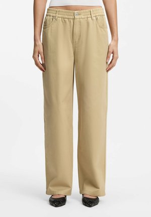 Kobieta nosząca beżowe spodnie o szerokich nogawkach z elastycznym paskiem i zapięciem na guzik, w połączeniu z czarnymi balerinami i białą bluzką.