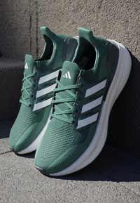 Zelené sportovní boty s texturovaným síťovaným svrškem, třemi bílými pruhy a polstrovanou bílou mezipodešví. Šnúrky jsou zelené, ladí s botou.